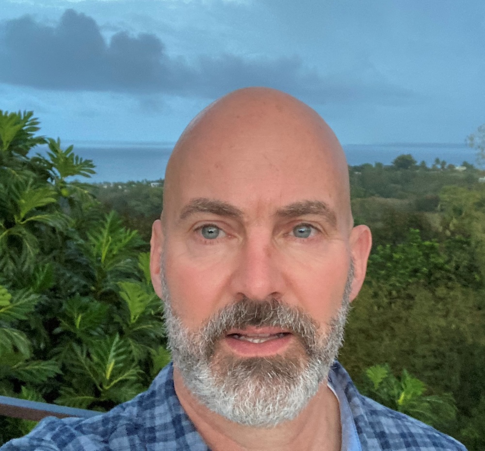 Bald man with a grey beard and blue eyes outdoors with a coastal background.